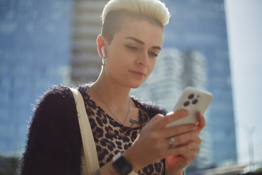 Portrait of a beautiful diverse woman with short blonde hair typing a message on smar phone and listening to music outdoor