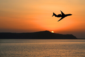 Silhouette of a private executive jet flying over an island at sunset. Luxury travel concept. Copy space. No people. - obrazy, fototapety, plakaty