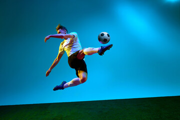 One sportsman, soccer player wearing yellow and black uniform hitting the ball in the air over football field in neon light. Concept of action, energy, sport