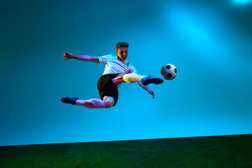 Professional man, football player in sports team uniform kicking the ball in motion over soccer field background in neon light. Scoring a goal.