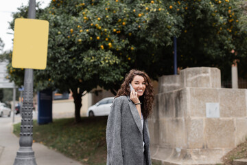 Fototapeta premium Carefree young curly woman in stylish casual jacket looking at camera while talking on smartphone and standing on urban street with street lamp and trees in Barcelona, Spain, at daytime
