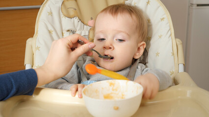 Fototapeta premium Closeup of mother feeding porridge to her baby son with spoon. Concept of parenting, healthy nutrition and baby feeding.