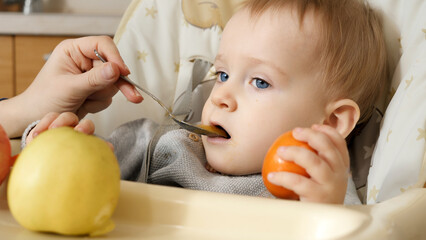 Cute baby boy sitting in highchair and eating porridge with fresh fruits. Concept of parenting,...