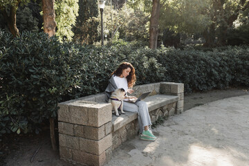 Fototapeta premium Curly freelancer in casual clothes using gadgets while spending time and working near pug dog sitting on stone bench in green park at daytime in Barcelona, Spain