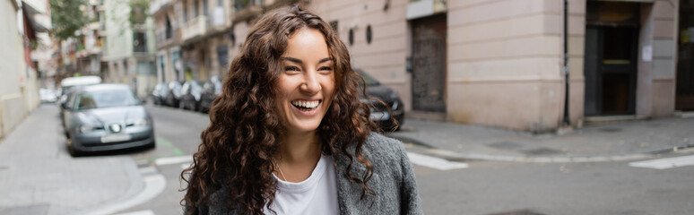 Cheerful and curly young woman in casual clothes and jacket looking away while standing on blurred urban street with buildings and cars at background in Barcelona, Spain, banner