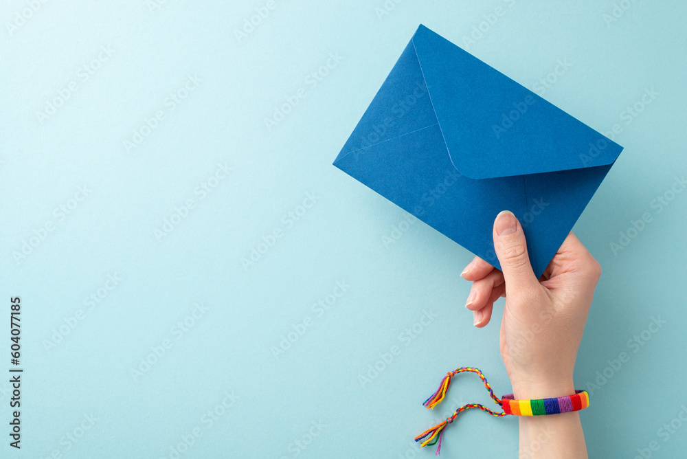 Wall mural LGBT History Month concept. First person top view photo of young person's hand wearing symbolic rainbow colored bracelet holding envelope on pastel blue background with space for text or advert