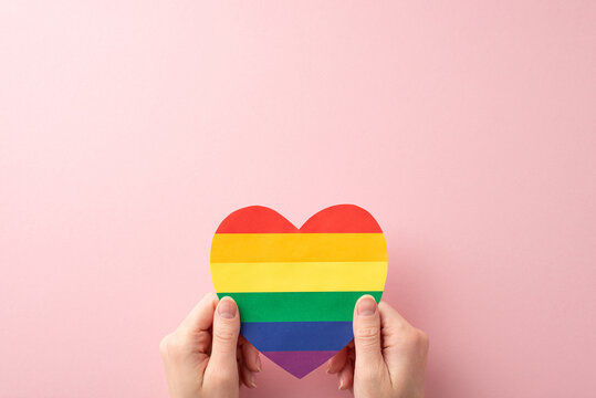 Join the celebration of LGBT History Month with this first person top view empowering image featuring a rainbow heart-shaped card held by woman's hands on pastel pink background