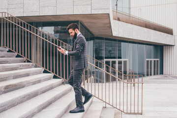 Young bearded elegant businessman going upstairs walking outdoors using smartphone