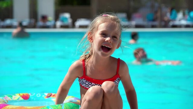 Little girl rests by hotel pool with blue water and inflatable swim ring and smiles. Child plays by water. Summer vacation, holiday and travel concept