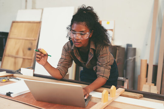 Female carpenter working with laptop computer and contact to customer in wood workshop. Female joiner wearing safety uniform and working in furniture workshop