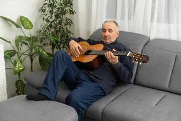 Senior man is playing guitar. Elderly man sitting on the sofa and playing guitar. Portrait of a gray-haired mature man in a sweater learning to play. Enjoying retirement life at home.