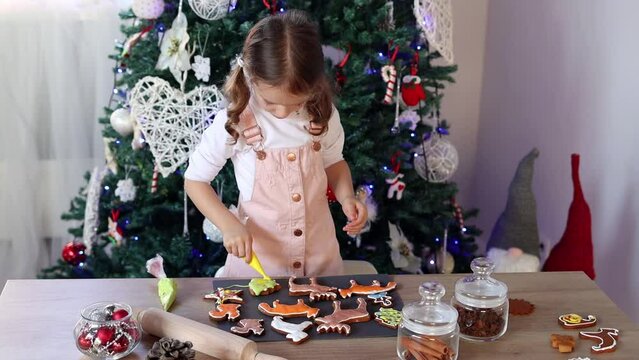 Happy little girl coloring Christmas gingerbread cookies at home in slow motion. Christmas decoration in the kitchen. Fir tree with fairy lights. The concept of the New Year and Christmas