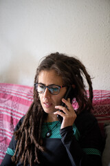 Woman with dreadlocks, glasses and piercings in her forties, Caucasian, talking on the phone on the sofa in her house with colourful covers.
