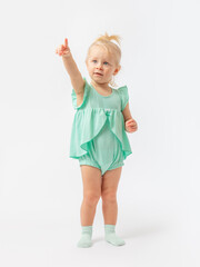 A 2-year-old toddler girl stands and seriously points her finger up against a white background in a green bodysuit dress and socks. Learning, playing.