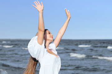 Fototapeta na wymiar Happy smiling beautiful woman on the ocean beach standing in a white summer dress, raising hands.