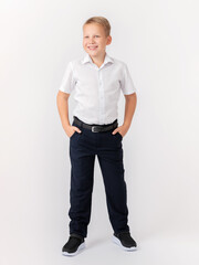 Cute confident teenager 10 years old blonde stands with his legs apart in a classic white shirt and dark trousers with a belt on a white background. Hands in pockets. Layout, advertising.