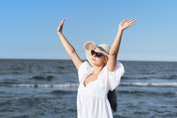 Happy blonde woman is on the ocean beach in a white dress and sunglasses, raising hands.