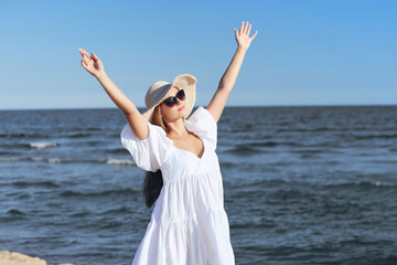 Fototapeta na wymiar Happy blonde woman is on the ocean beach in a white dress and sunglasses, raising hands.