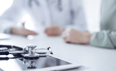 Doctor and patient are sitting and discussing something at the desk in the clinic office. The focus is on the stethoscope lying on the table, close up. Perfect medical service and medicine concept.