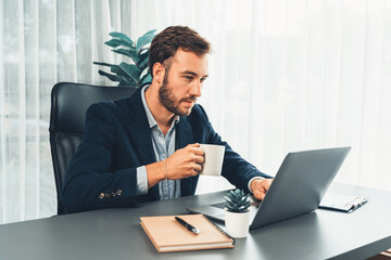 Businessman in black suit working on laptop at his workspace desk. Smart executive researching financial data and planning marketing strategy on corporate laptop at modern workplace. Entity