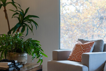 Detail view of living room chair with decorative pillow near potted plants and large window view of autumn foliage.