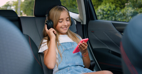 Small stylish girl smiling riding in car, listening to music with headphones on smartphone in backseat of car.