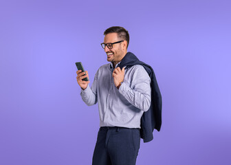 Happy young businessman holding blazer and checking messages on smart phone. Male professional entrepreneur smiling and scrolling social media while standing isolated against blue background