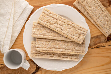 Several rye crispbreads with white plate, cup and linen white napkin on wooden table, macro, top view.