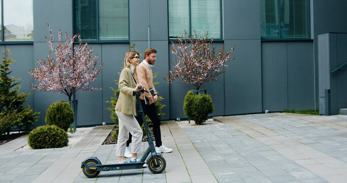 Caucasian young woman and man walking on street with kick e-scooter and laptop talking and laughing by modern office buildings. Technology, partnership, outside