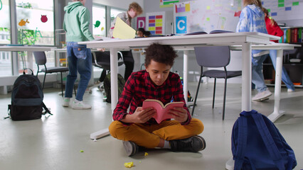 African-American teen student read book sitting under desk in noisy class