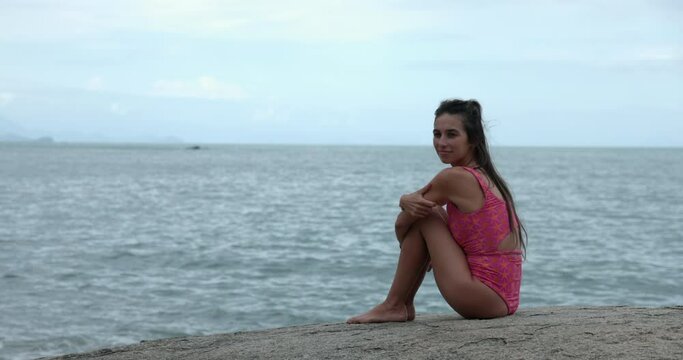 Woman sitting on rocks near ocean sad looks back towards camera - wide shot