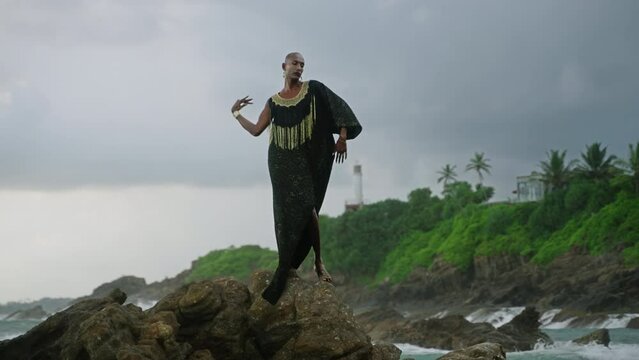 Queer black person gracefully poses in posh dress, handmade jewelry stands on scenic sea cliff top above dramatic cloudy sky, ocean waves. Lgbtq biethnic gay model in brass accessories.