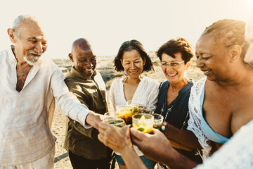 Happy multiracial senior friends having fun drinking and toasting mojitos on the beach during sunset time - Elderly people enjoying summer holidays