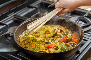 A woman's hand puts udon in a frying pan with stewed vegetables in spices.