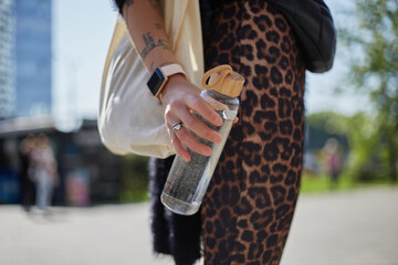 Young woman holds a glass bottle of water in hand. Responsible female person wearing cotton shopper bag on shoulder and holding a reusable water bottle