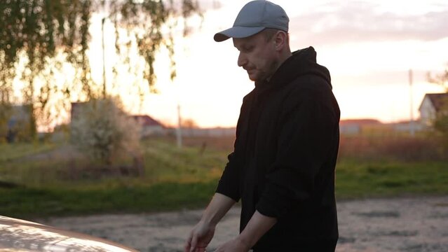Young man is opening the hood of a car outdoors in the evening on sunset. Driver checking or fixing the automobile engine on the road after a car breakdown. Vehicle service concept. Slow motion.