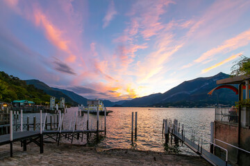 Fototapeta na wymiar A ferry pulls away from the Lungolago Europa promenade as the sun sets and the sky turns pink over the mountains and Lake Como, in Bellagio, Italy.