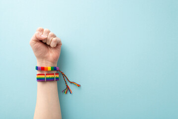 A raised hand wearing a rainbow-colored wristlets with a clenched fist symbolizing strength and the fight for rights takes center stage in this photo on pastel blue backdrop with space for text or ad - obrazy, fototapety, plakaty