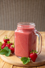 Strawberry smoothie or milkshake on wooden board with fresh strawberry fruit and green leaves. Strawberry drink in jar mug full of vitamins and antioxidants.