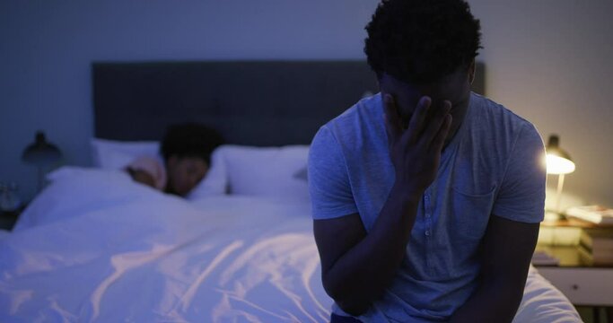 Infertility, depression and a black man sitting on the bed with his wife sleeping at night. Erectile dysfunction, infidelity and sleepless with a husband feeling sad in the bedroom of his home
