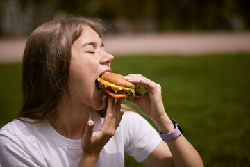 a young girl takes out a burger from a paper bag sitting on the green grass, the concept of food delivery, street food