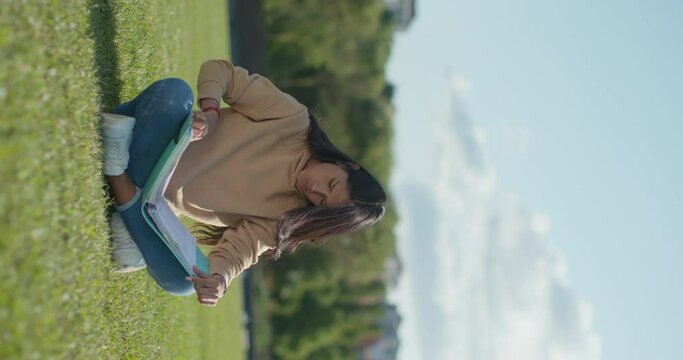 Young teenage Latina college student studying for an exam in an outdoor park with books, vertical shot