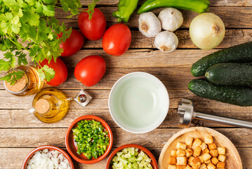Necessary ingredients to make a gazpacho around an empty bowl, and with the chopped vegetables and the toasted bread to pour it on top, on a rustic wooden table, top view.