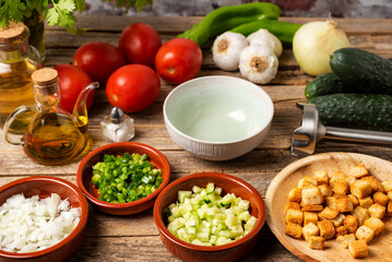 Necessary ingredients to make a gazpacho around an empty bowl, and with the chopped vegetables and toasted bread to pour it on top, on a rustic wooden table.