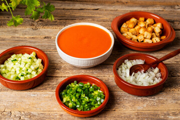 Bowl with gazpacho next to some clay plates with chopped vegetables and fried bread to add to the gazpacho, on a rustic table.