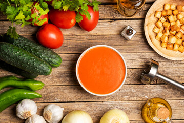 Vegetables needed to make a gazpacho around a bowl filled with gazpacho and chunks of toasted bread, on a rustic wooden table, top view.