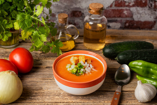 Vegetables necessary to make a gazpacho around a bowl full of gazpacho with chopped vegetables and chunks of toasted bread, on a rustic wooden table, diner view.