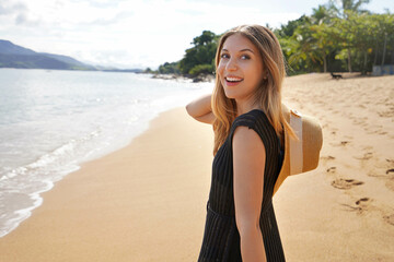 Beautiful slim girl with black sundress walking on the beach carrying straw hat on her shoulder turns behind and smile. Summer holidays and relax on the beach.