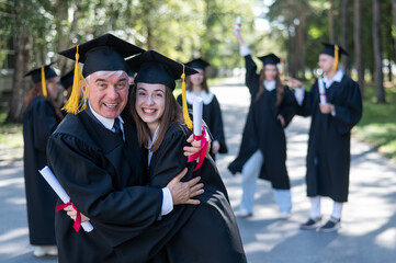 A group of graduates in robes outdoors. An elderly man and a young woman congratulate each other on...