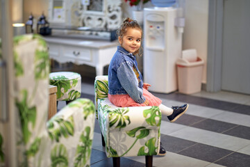 Adorable toddler girl sitting on modern upholstered chair in a furniture shop, looking around.